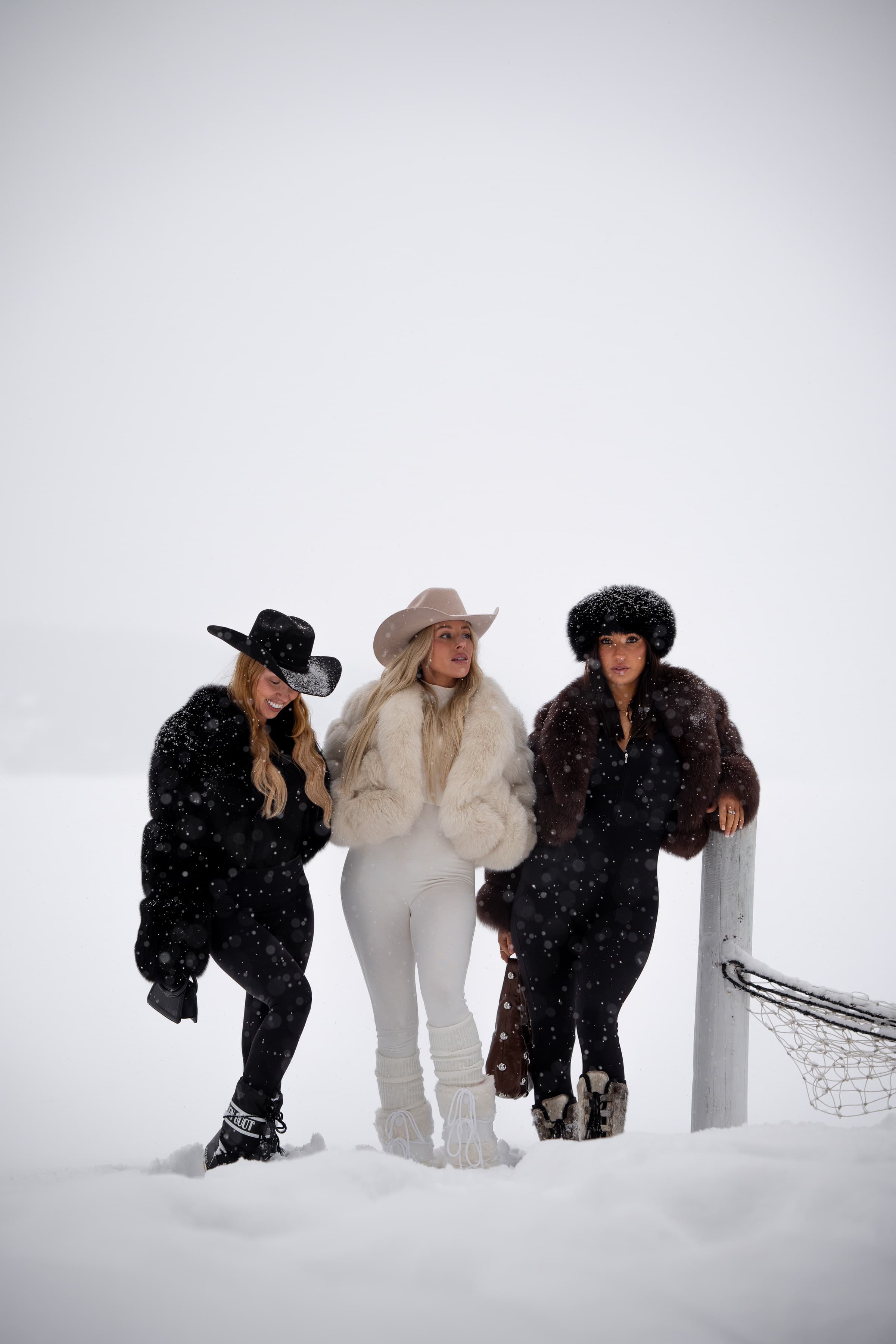 Three FARAYA fox coats — black, ivory and brown — worn together in falling snow.
